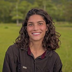 Zhane Moledina has shoulder-length wavy brown hair and wears a brown collared shirt while standing in front of a green landscape.